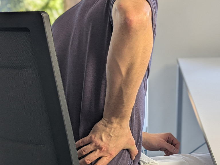 Mann sitzt auf Stuhl und hält Hand an unteren Rücken
